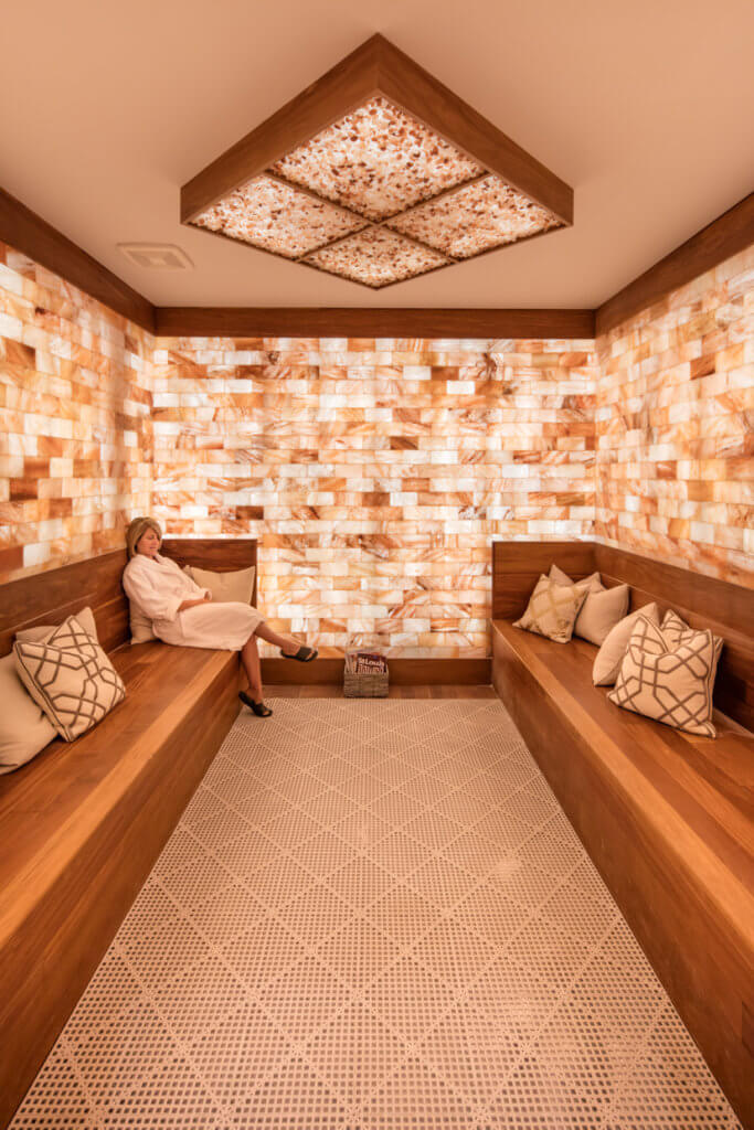 PLAM Health. Luxurious spa room with two, brown benches facing each other. The room's walls are completely tiled. On the ceiling, is a large, squared formation full of salt rocks. Sitting on one of the benches is a woman in a white robe.