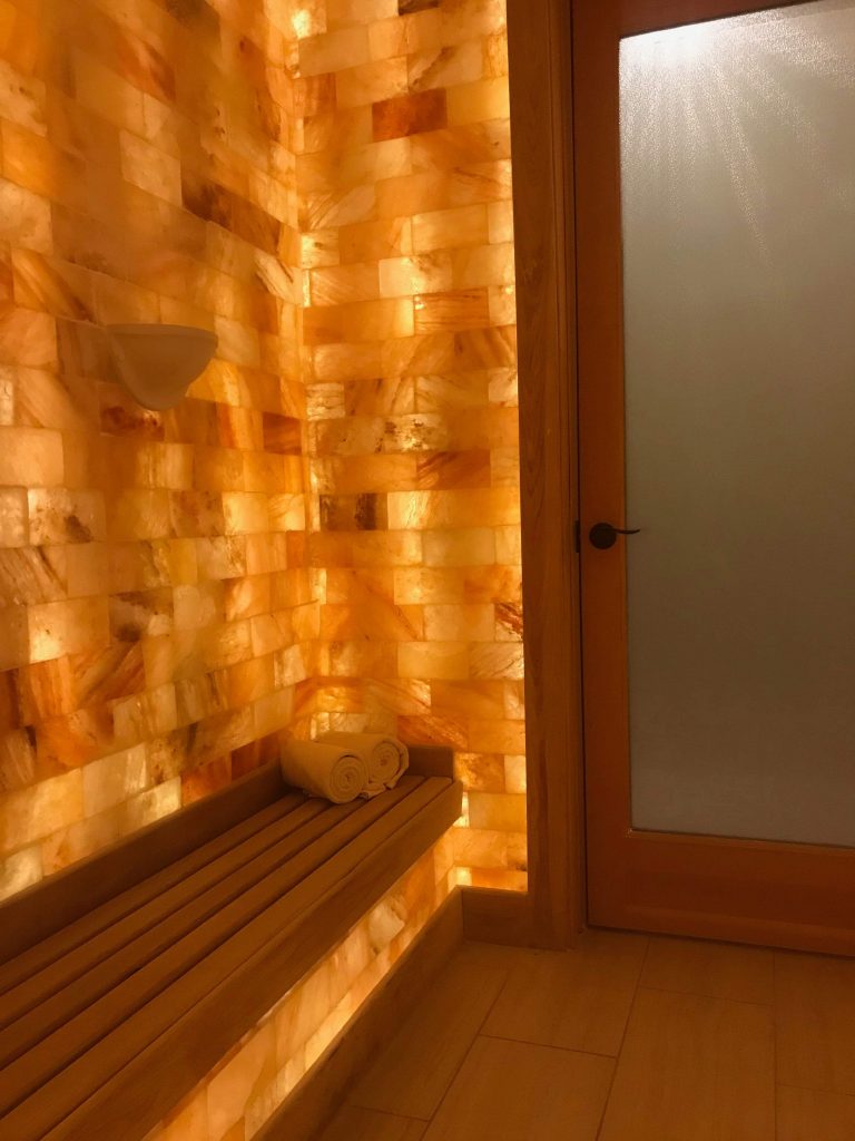 The Henderson Beach Resort &Amp; Spa. Narrow Spa Room With Wooden Bench Wrapped Around The Room. All Of The Walls Are Very Light Colored Salt Tiles. View From Back Of Room Facing The Door.