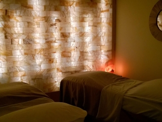 Two Massage Tables With Pillows And Blankets In A Small Room With A Led Backlit Salt Brick Wall. There Is A Round Mirror Hanging On The Wall To The Right, And A Lit Himalayan Salt Lamp Propped Up On A Table In The Corner Of The Room.
