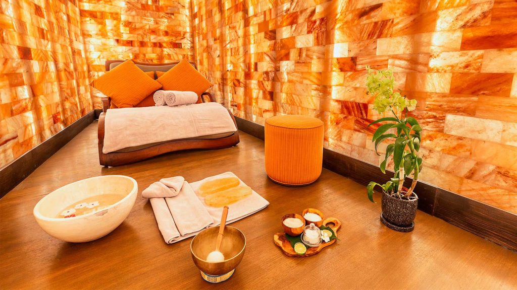 Sulha Spa At Kudadoo. Large Spa Room With A Bed Placed In The Middle. In Front Of The Bed, Are Bowls Of Different Natural Treatments And Remedies, There Is Also A Plant As Well As A Towel Placed On The Floor.