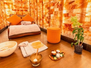 Sulha Spa At Kudadoo. Large Spa Room With A Bed Placed In The Middle. In Front Of The Bed, Are Bowls Of Different Natural Treatments And Remedies, There Is Also A Plant As Well As A Towel Placed On The Floor.