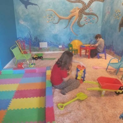 Young girl sitting on the ground and boy sitting at a red table playing with toys in a salt-covered floor playpen with an ocean painted wall