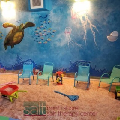 Four small chairs on a salt-covered floor with an ocean painted wall and "Salt north shore salt therapy" on the bottom of the picture.