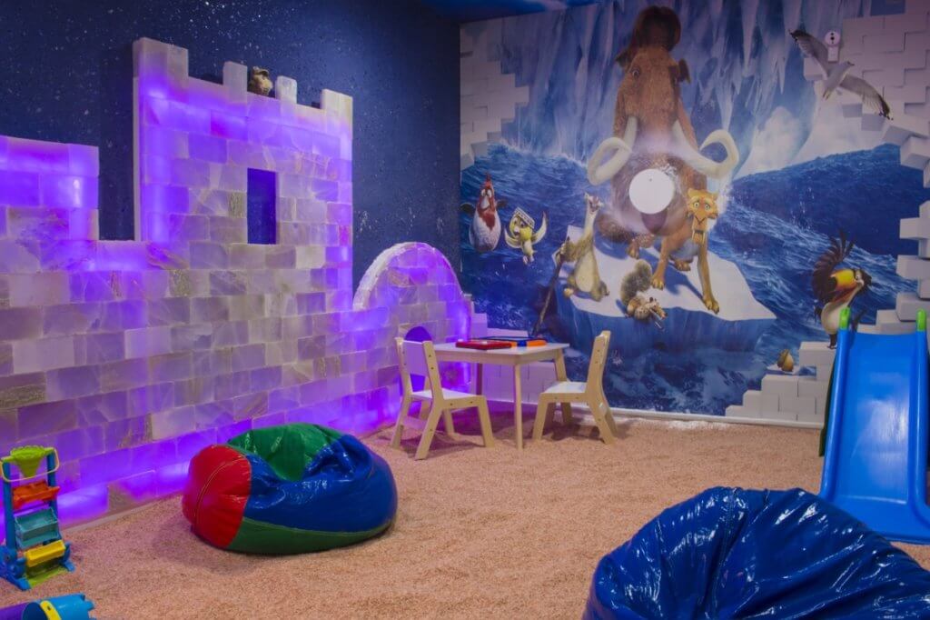 Salt Breeze. Large salt room made for children. Back wall is painted as an ice age mural and put together on the side wall are salt blocks in the shape of a castle. On top of the salted floor are bean bags and other toys.