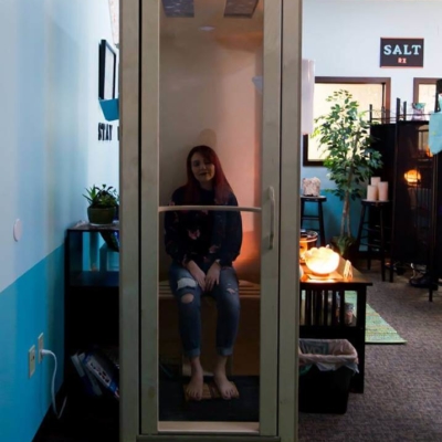 Woman relaxing in a light wooden and glass salt booth surrounded by white and blue walls and decor at the Peace of Salt in Everett, Washington