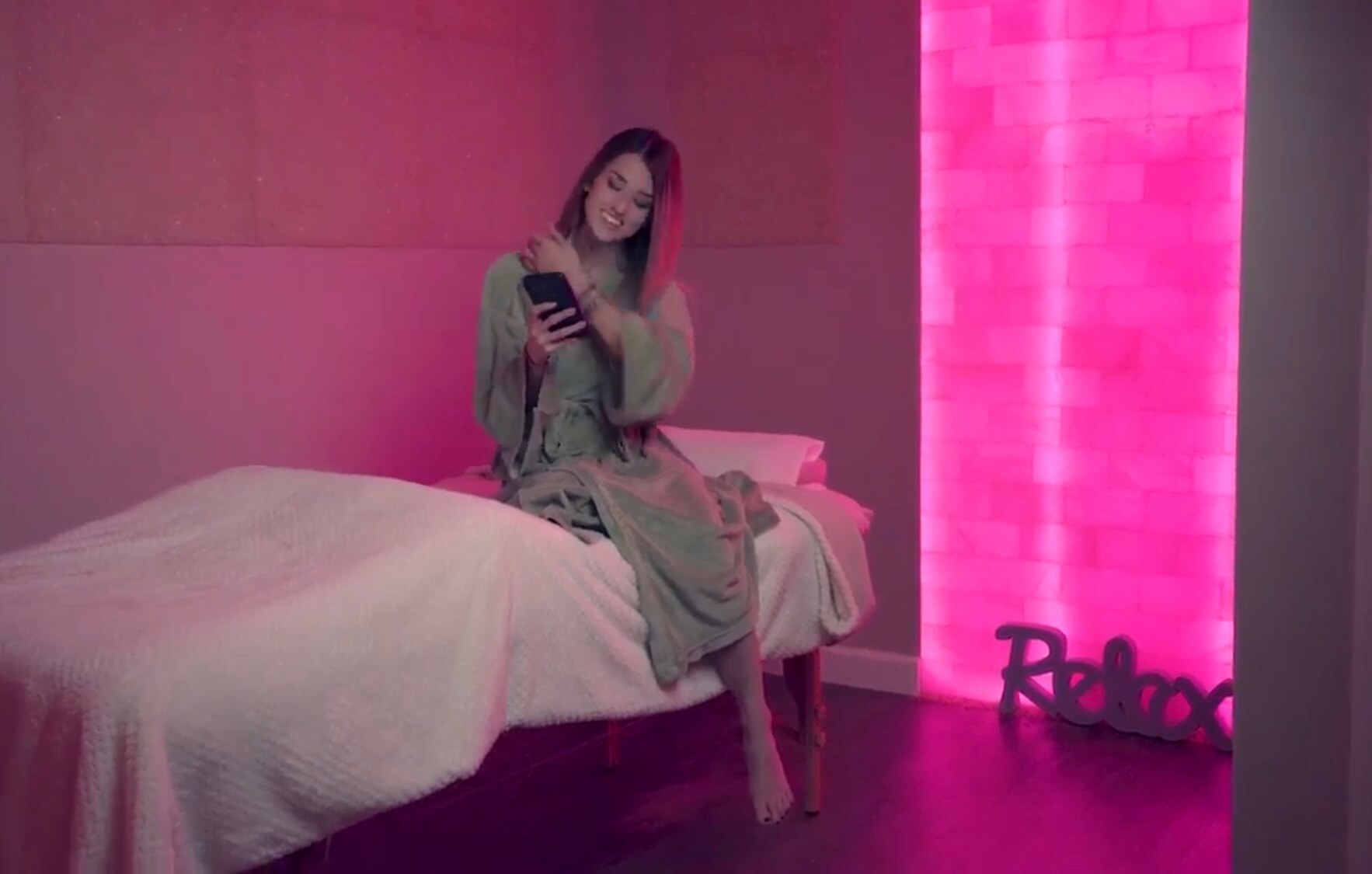 Woman in a green robe sitting on a cushioned table covered by a white blanket with a pink backlit salt stone wall and square salt stone decor at the Premier Day Med Spa in Hallandale Beach, Florida