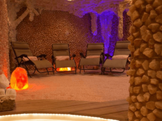 Rain Wellness Spa. Salt Room With 4 Lounge Chairs In It. Walls Are Old Fashioned, Cave-Like Rock Formations With A Glowing Purple Light In The Back Right Corner. Scattered Around The Room Are Glowing Salt Rocks.