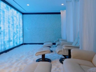 Salt. Large, Salt Room Illuminated With Blue Lights, Has 4 Chairs (Each With An Ottoman) Sitting On Top Of The Salt Covered Floor. Between Each Chair Is A White Curtain That Can Be Used For Privacy.
