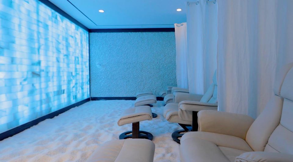 Salt. Large, salt room illuminated with blue lights, has 4 chairs (each with an ottoman) sitting on top of the salt covered floor. Between each chair is a white curtain that can be used for privacy.