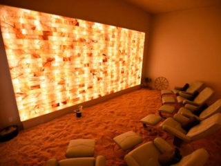 Saltbox Dry Salt Therapy. Numerous Chairs And Ottomans Placed Next To Each Other In Salt Room Facing Large, Illuminated Salt Wall.