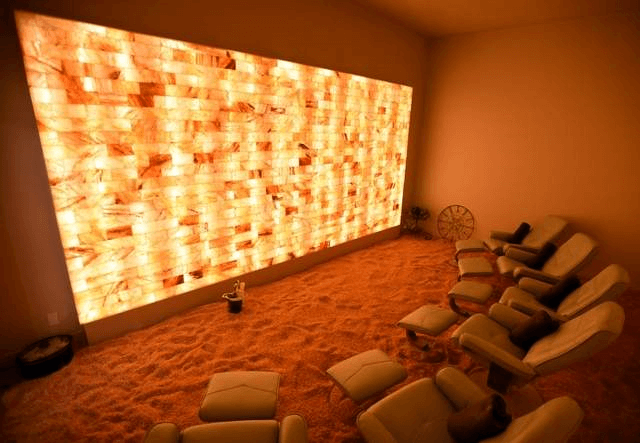 Saltbox Dry Salt Therapy. Numerous chairs and ottomans placed next to each other in salt room facing large, illuminated salt wall.