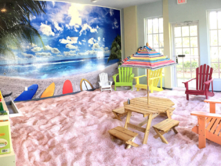 The Salt Box. Large, beach themed salt room. Large image of the beach grabs the attention in the front of the room with adirondack chairs scattered around on top of the salt covered floor to represent sand.