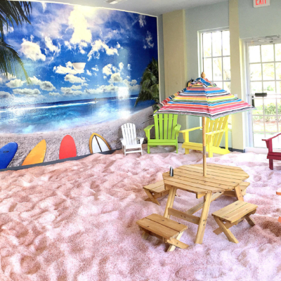 The Salt Box. Large, beach themed salt room. Large image of the beach grabs the attention in the front of the room with adirondack chairs scattered around on top of the salt covered floor to represent sand.