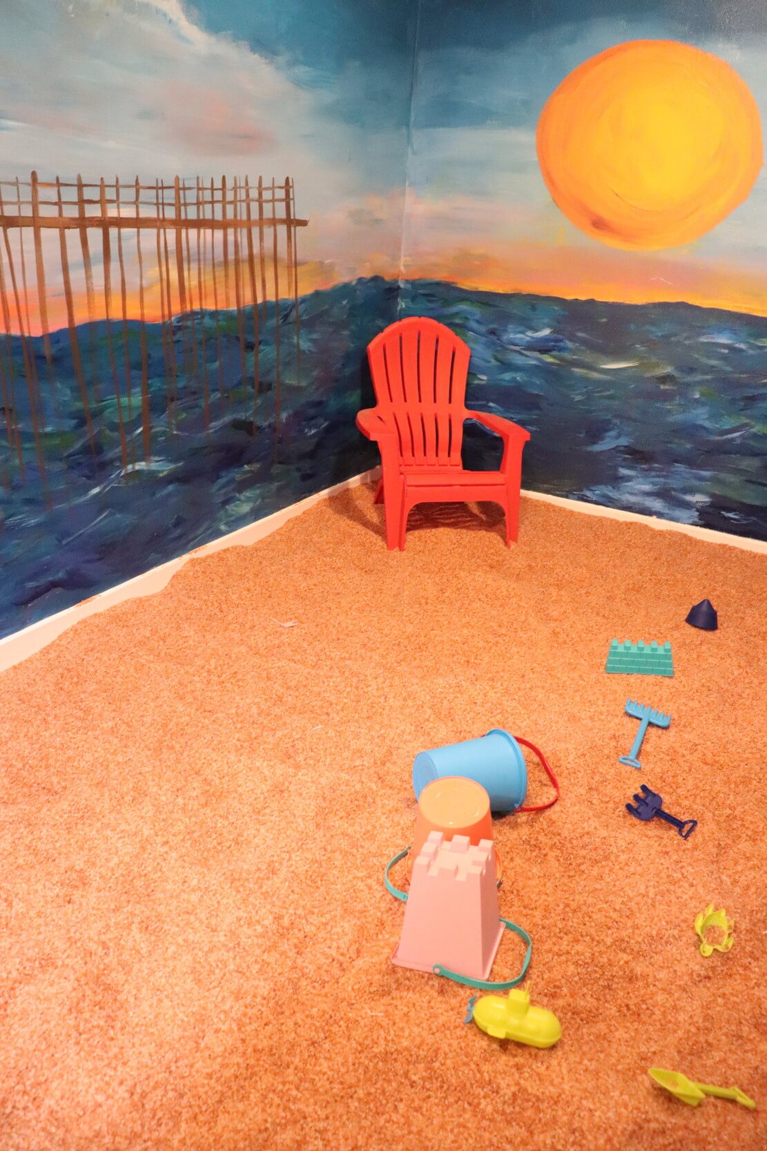 A sunset on the ocean painted wall with a red chair on a Himalayan salt-covered floor with children's toys on the floor.