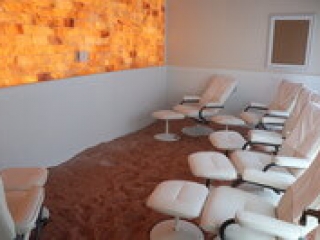 Salt Life Spa. Small Salt Room With 6 White Chairs And Ottomans Scattered Around The Room Facing A Salt Wall.
