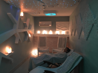 Sebring Salt Spa. Small, Dimly Lit Salt Room With A Woman Laying Back Sleeping In Recliner. Room Is Illuminated By Glowing Salt Rocks And Lights On The Ceiling Meant To Represent Stars.