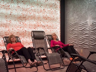 The Acupuncture Center And Salt Spa. Two Women Sit In Lounge Chairs With A Large Salt, Stone Wall Behind Them. One Woman Is In A Salmon Colored Shirt While The Other Is In A Magenta Shirt.