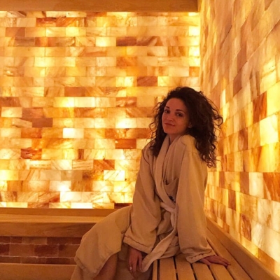 Woman in a robe sitting on a wooden bench in a salt room surrounded by LED backlit salt panel walls at The Ritz Carlton Spa - Charlotte, North Carolina.