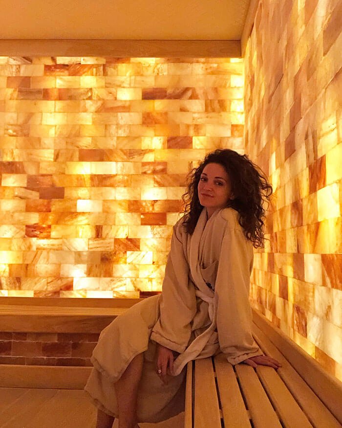 Woman In A Robe Sitting On A Wooden Bench In A Salt Room Surrounded By Led Backlit Salt Panel Walls At The Ritz Carlton Spa - Charlotte, North Carolina.