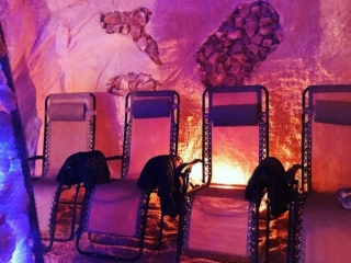 The Salt Cave. Lounge Chairs Lined Up In Salt Cave With Towels Draped Over Them. Cave Is Glowing With Pink And Blue Lights And Salt Is Covering The Whole Floor.