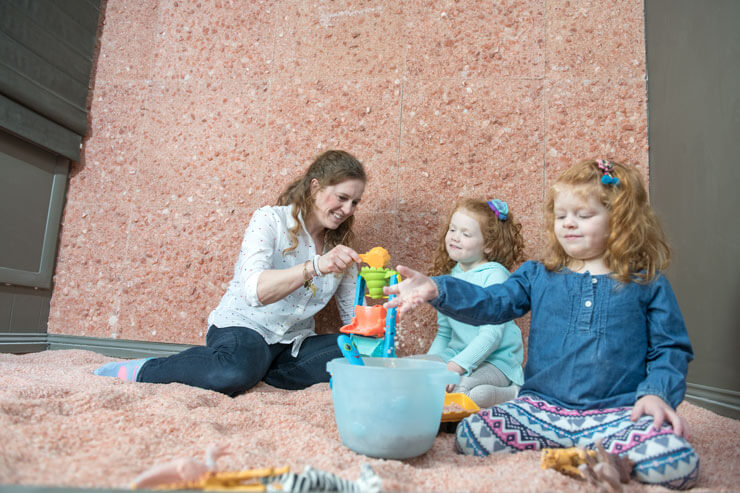A women and two young girls sitting and playing a salt-covered floor in front of a Himalayan salt stone wall.