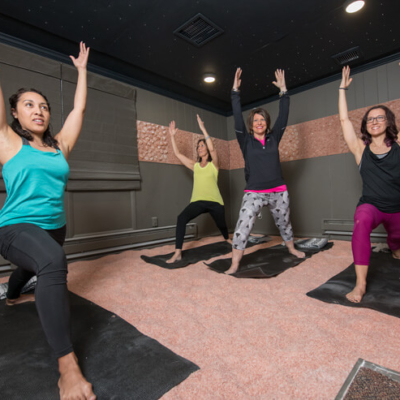 Four Women On Black Yoga Mats In Warrior One Doing Yoga In A Salt Therapy Room With A Salt-Covered Floor And A Salt Stone Center Panel All The Way Around The Wall