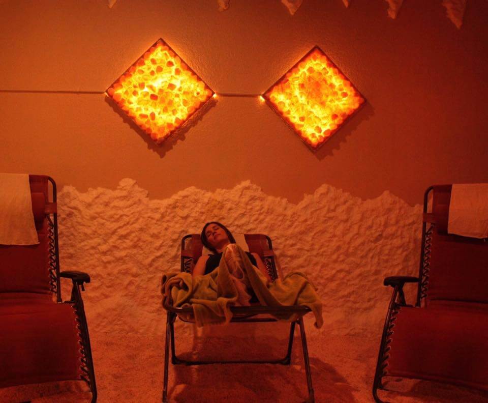 Three Reclining Chairs With The Women Relaxed In The Center Chair Under A Blanket In A Salt Studio With Two Diamond Salt Stone Décor Black Lit By Orange Lights.