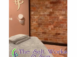 The Salt Works And Spa. Massage Bed With Towels On It. Focus Is On The Salt Tiled Wall And Logo Mounted On The Wall. Text Over Image Reads &Quot;The Salt Works &Amp; Spa Halotherapy - Wellness - Beauty&Quot;