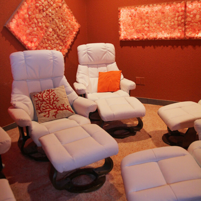 Two White Cushioned Chairs With Pillows And Foot Rests In A Salt Therapy Room On A Salt-Covered Ground With Himalayan Salt Stone Décor.