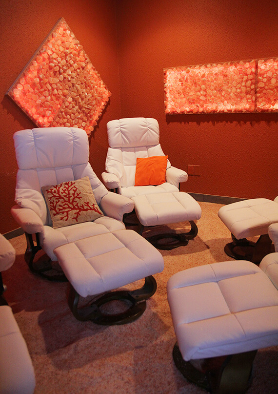 Two White Cushioned Chairs With Pillows And Foot Rests In A Salt Therapy Room On A Salt-Covered Ground With Himalayan Salt Stone Décor.