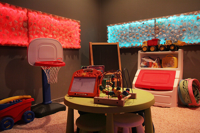 Children'S Play Room With Toys, A Mini Basketball Hoop, And A Table On A Salt-Covered Floor With One Red And One Blue Rectangular Backlit Himalayan Salt Décor On The Walls..