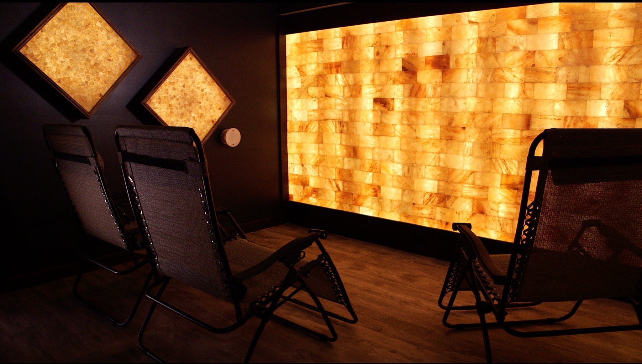 Three reclining chairs on a brown wooden floor in front of an orange backlit salt wall with two diamond orange backlit salt stone decor.