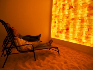 The Salt Spa. A Woman And Young Girl Lay Back In Lounge Chairs In A Salt Room And Observe The Large, Illuminated Salt Tile Wall In Front Of Them.