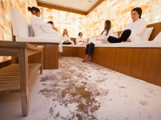 Six Women In White Robes Sitting On A Wooden Bench With White Cushions Relaxing In A Salt-Covered Floor Room With Salt Stone Walls Backlit By Orange Lighting