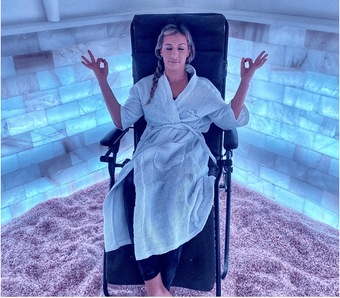 Salt & Sweat. Woman relaxes in lounge chair while meditating. While doing this, she is in the corner of a salt room with illuminated walls.