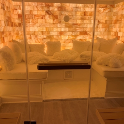A Halotherapy Room Through A Glass Door With A White Cushioned Bench With Pillows And A Backlit Himalayan Salt Stone Walls.