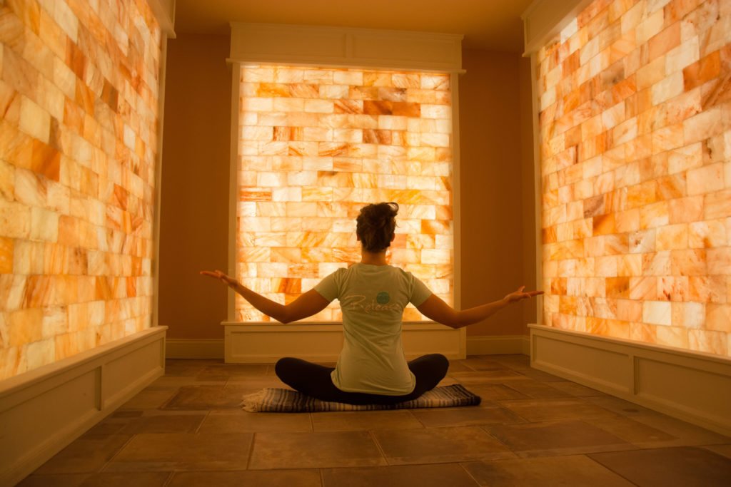 Release Well-Being Center. Woman is sitting and appears to be meditating inside of spa room surrounded by salt tiles on wall.