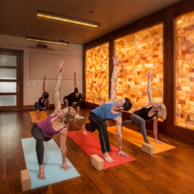 Two Men And Three Women All On Yoga Mats With Yoga Black In A Twist Position In Front Of A Salt Stone Paneled Wall Backlit By Orange Lighting.