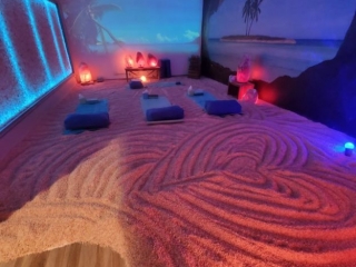 Salt Sensations Usa. Long, Colorful, Beach Themed Salt Room Illuminated By Blue Lights And Glowing Salt Rocks. In The Middle Of The Room Lay 4 Beds On Top Of The Salted Floor.