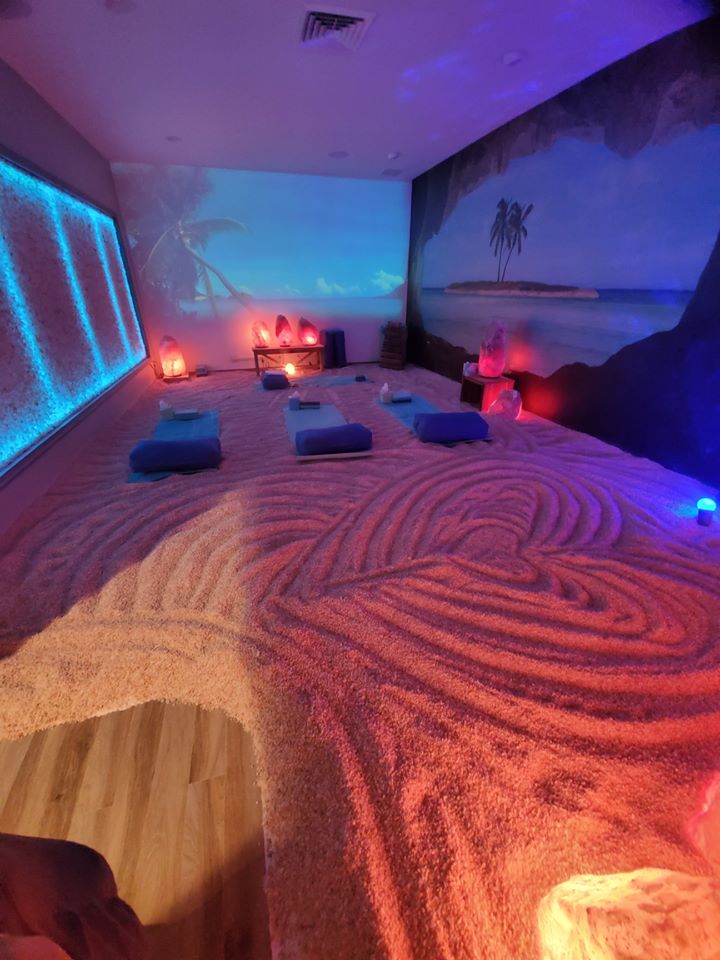 Salt Sensations USA. Long, colorful, beach themed salt room illuminated by blue lights and glowing salt rocks. In the middle of the room lay 4 beds on top of the salted floor.
