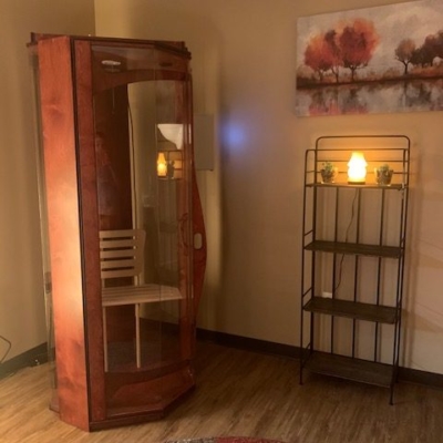 Wooden burgundy glass halotherapy booth in a tan room with grey vinyl flooring, a brown shelf with a painting above, and grey and red an area rug at Sole Purpose Foot Care in Florence, Kentucky