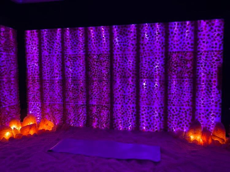 Pellegrino Healing Center. Large salt room completely lit up by purple lights. In the center of the salt room lays a yoga mat and in the corners of the room, sit glowing salt rocks.