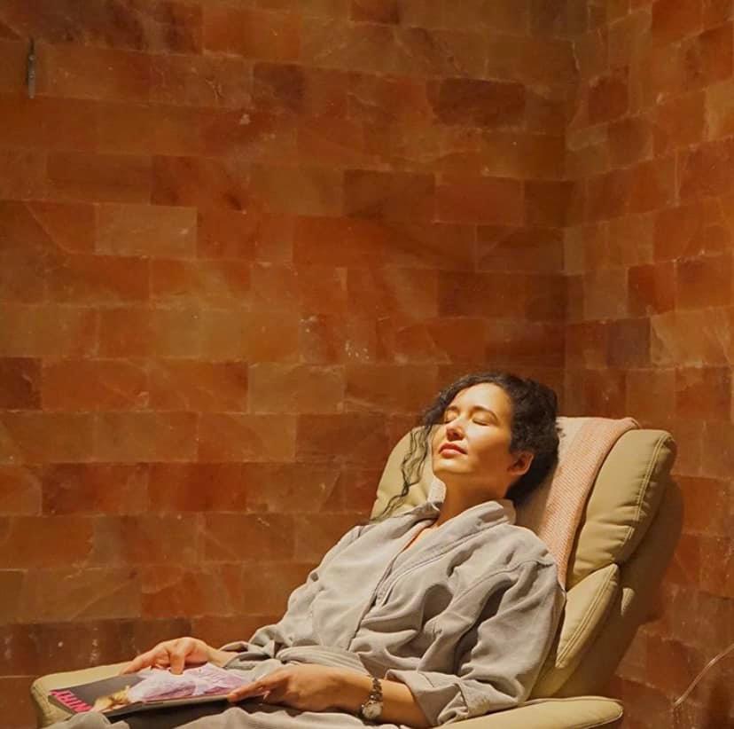 Art Of Balance Wellness Spa - Woman In Grey Robe Laying Back In Chair In Front Of A Himalayan Salt Brick Wall.