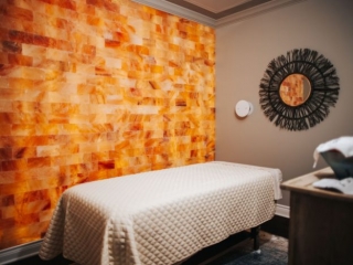 The Woodhouse Day Spa. Massage Bed With White Blanket Over It. Above The Bed, Mounted On The Wall Us A Sun-Shaped Mirror.