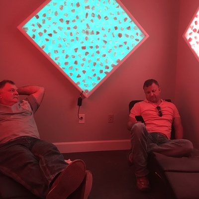 Two men, one relaxing and the other sitting on his phone, in a room with two diamond salt stone decor, one backlit by blue lighting and the other with pink lighting.