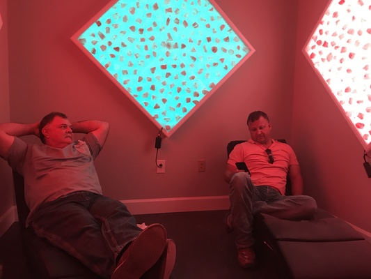 Two men, one relaxing and the other sitting on his phone, in a room with two diamond salt stone decor, one backlit by blue lighting and the other with pink lighting.