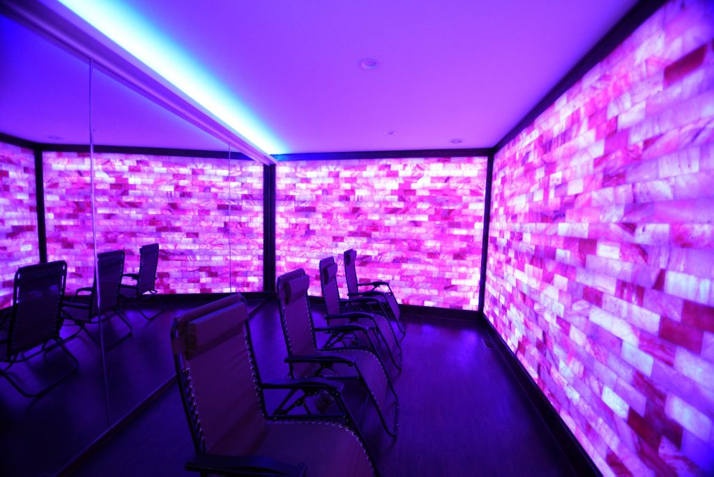 Float into Wellness and the Salt Lounge & Sauna Spa. Four lounge chairs in spa room lit by purple lights,