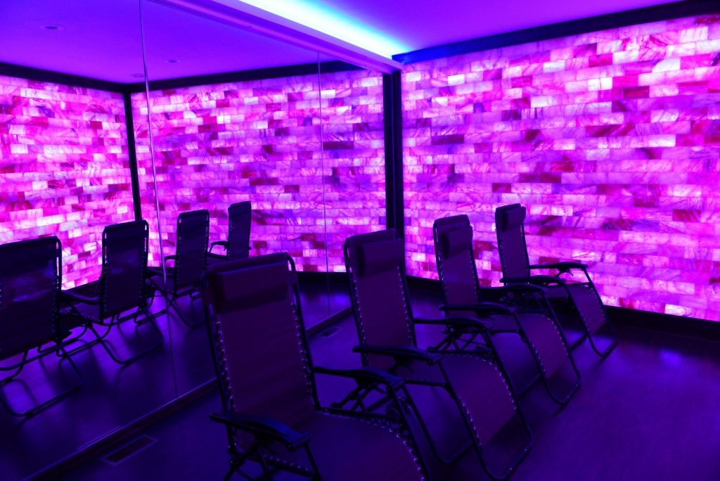 Float into Wellness and the Salt Lounge & Sauna Spa. Four lounge chairs in spa room lit by purple lights,
