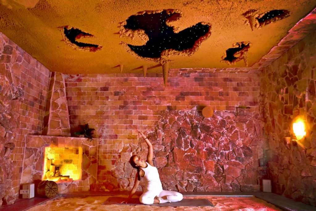 Healing Salt Cave & Wellness Spa. Woman is doing yoga inside of a salt room. The ceiling has a cave-like feel to it and the walls appear to be brick-like.