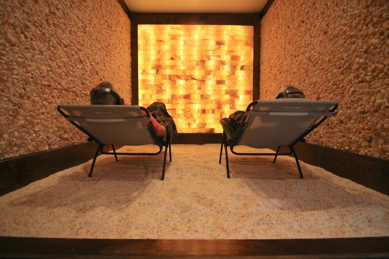 Two Men Relaxing In Lounge Chairs On A Bed Of Salt With Headsets On In A Salt Room With A Led Back Lit Salt Brick Wall And Two Salt Panel Walls At The Intown Salt Room - Atlanta (Grant Park), Georgia.